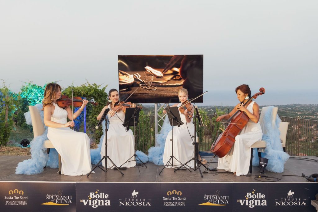 Quartetto d’archi in abito bianco si esibisce tra i vigneti dell’Etna durante l’edizione 2025 di Bolle in Vigna, evento promosso da Tenute Nicosia che unisce spumanti etnei, musica e paesaggio.