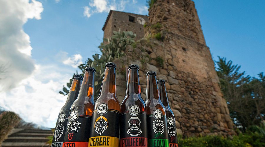 Bottiglie di birra artigianale Epica davanti a una torre in pietra in Sicilia, simbolo del legame tra produzione brassicola contemporanea e territorio storico.