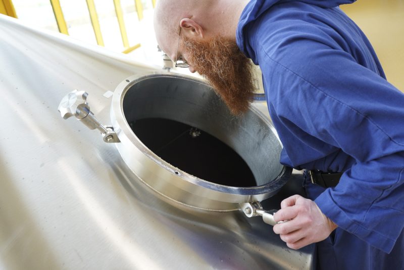 Monaco in abito blu che controlla la lavorazione della birra all’interno di un birrificio monastico, simbolo della tradizione brassicola dei monasteri europei.