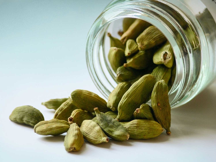 Baccelli di cardamomo verde intero fuoriusciti da un barattolo di vetro, spezia aromatica dalle origini orientali utilizzata nella cucina e nella cultura mediterranea.